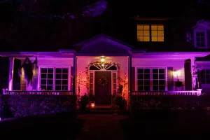Front view of a house decorated for Halloween at night, illuminated with purple and orange landscape lighting. A skeleton and grim reaper decoration hang on the porch, with pumpkins and festive lights around the entryway.