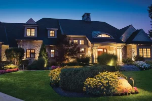 A large residential home at dusk with professional outdoor lighting highlighting the front entrance, stone facade, garden beds, trees, and walkway for enhanced curb appeal and nighttime visibility.