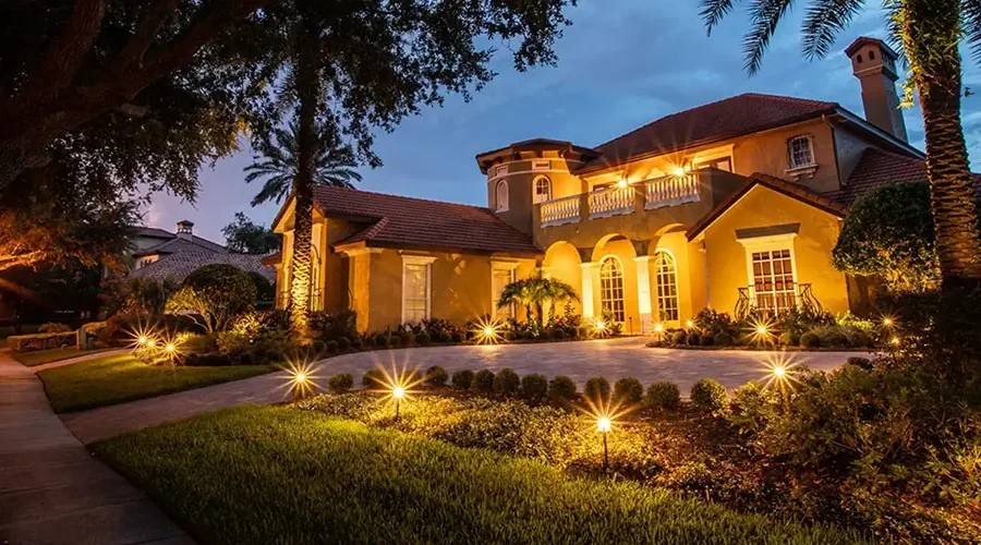Home exterior lit with warm yellow landscape lighting along pathways and around trees, creating a welcoming glow at dusk.