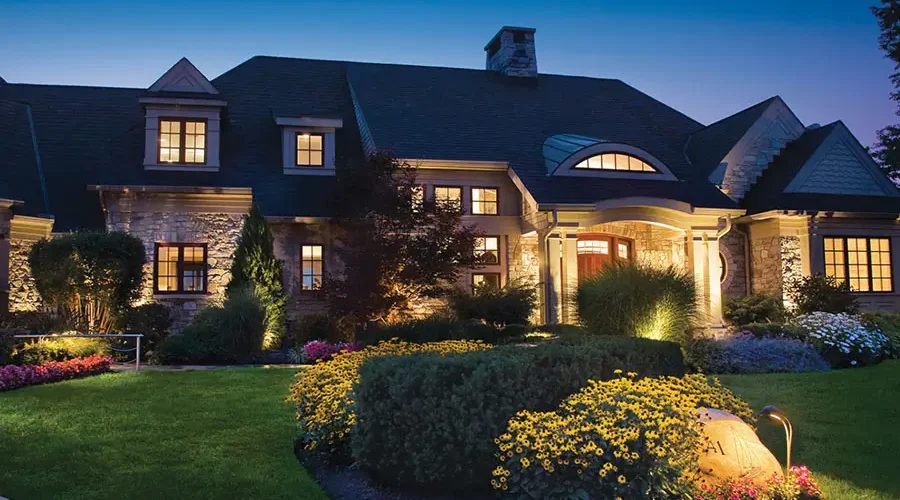 A large residential home at dusk with professional outdoor lighting highlighting the front entrance, stone facade, garden beds, trees, and walkway for enhanced curb appeal and nighttime visibility.