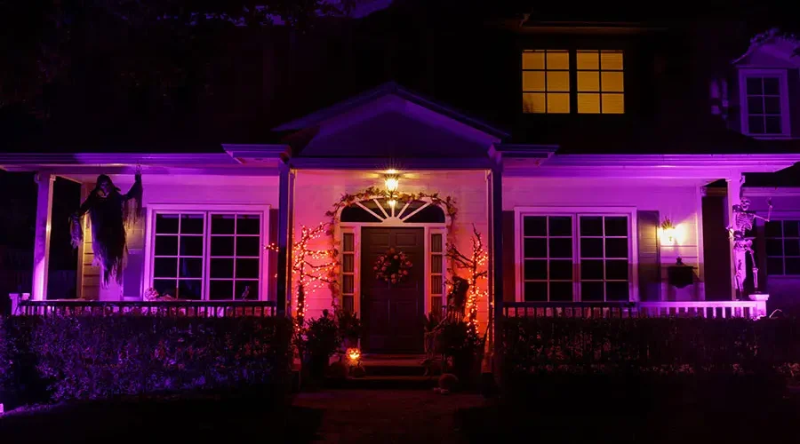 Front view of a house decorated for Halloween at night, illuminated with purple and orange landscape lighting. A skeleton and grim reaper decoration hang on the porch, with pumpkins and festive lights around the entryway.